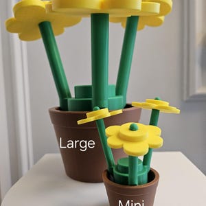 May include: Two 3D printed flower pots, one labeled "Large" and the other labeled "Mini". The larger pot has three yellow flowers with green stems. The smaller pot has two yellow flowers with green stems.