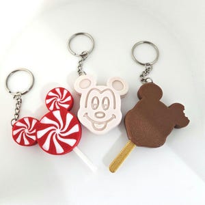 May include: Three keychains featuring a red and white peppermint candy, a white Mickey Mouse head, and a brown chocolate ice cream bar. Each keychain has a silver metal ring and chain.
