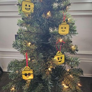 May include: A Christmas tree decorated with four yellow Lego minifigure ornaments. The ornaments are hanging from red ribbon.