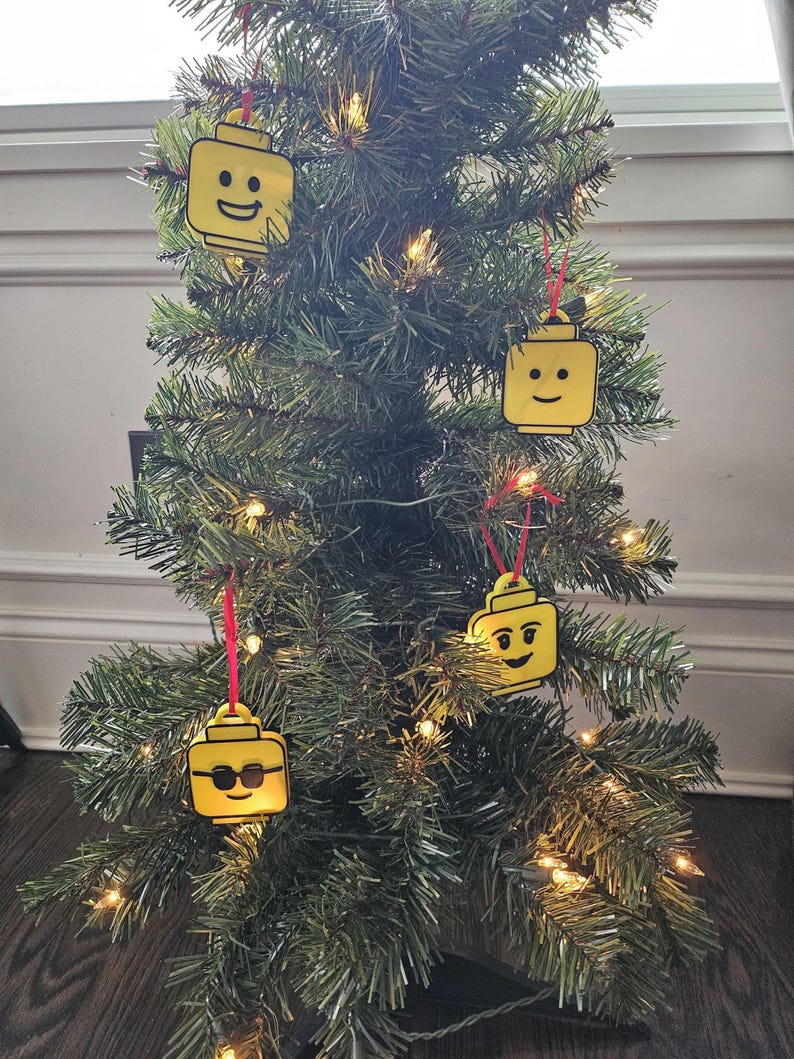 May include: A Christmas tree decorated with four yellow Lego minifigure ornaments. The ornaments are hanging from red ribbon.