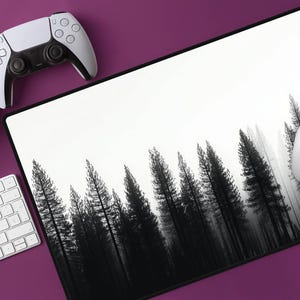Puede incluir: Un tapete de ratón para juegos blanco y negro con un diseño de silueta de bosque en blanco y negro. Un mando inalámbrico blanco y un ratón inalámbrico blanco están sobre una superficie morada.