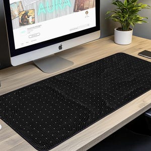 Könnte beinhalten: Eine schwarze Schreibtischunterlage mit einem dezenten Gittermuster und weißen Punkten. Die Unterlage liegt auf einem Holztisch mit einem Computermonitor, einer Topfpflanze und einem Notizbuch.