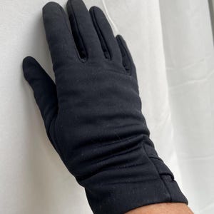May include: A black glove made of a soft, stretchy fabric. The glove is designed to fit the hand snugly, with a gathered detail at the wrist. The fabric appears to be a solid, matte black.