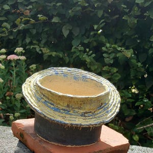Puede incluir: Una maceta cilíndrica de cerámica con un borde texturizado y superpuesto que presenta un acabado desgastado en amarillo y azul. La base del recipiente es de un color gris oscuro sólido, contrastando con la sección superior e interior más claras.
