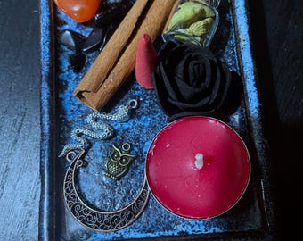 May include: A blue ceramic tray containing a selection of items, including a cinnamon stick, a small glass jar with green herbs, a red candle, a black rose, a small orange crystal, a silver snake charm, and a gold owl charm.