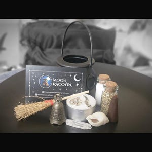 May include: A black cauldron, a small broom, a metal bell, a clear crystal, a seashell, two glass vials with cork stoppers, and a round candle with a pentagram charm. A black business card displays the text Moon Racoon with a raccoon illustration.