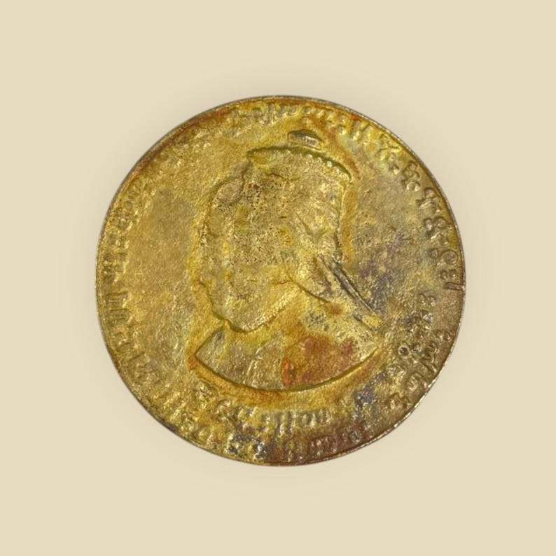 May include: A gold-colored coin with a profile of a woman wearing a crown on the obverse. The coin has a raised rim and is slightly worn.