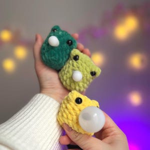 May include: Three handmade crochet frog toys in green, yellow, and light green. Each frog has black eyes and a white bubble. The frogs are held in a person's hand, with a soft, white sweater in the background.