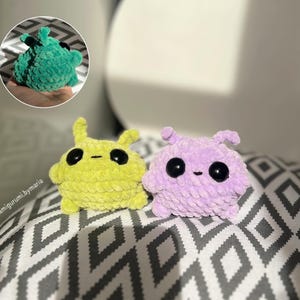 May include: Two handmade, plush amigurumi creatures in the image. One is lime green, the other is lavender. Both have large black eyes and small, rounded ears. A smaller, green amigurumi is in a circular inset. The background is a patterned fabric.