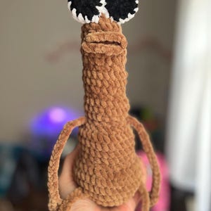 Può includere: Una creatura all'uncinetto marrone fatta a mano con grandi occhi neri e bianchi. Il peluche ha un corpo lungo e strutturato e piccole braccia e gambe. Il design stravagante è morbido e coccoloso.