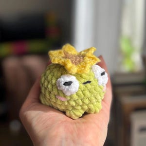 May include: A handmade, crocheted frog with a yellow sunflower on its head. The frog is light green with white eyes, black pupils, and pink cheeks. The sunflower has yellow petals and a brown center. The frog is small and appears to be soft.