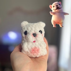 May include: A small, white, plush, crocheted animal figure with black plastic eyes, a black nose, and pink accents on the cheeks and belly. Floating above the plush is a small, digital, light-pink, cartoon-style French bulldog puppy illustration.
