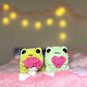 May include: Two handmade crochet frog plushies. One is lime green holding a pink flower bouquet, the other is mint green with a pink heart. Both have black eyes and rosy cheeks, set on a pink blanket.