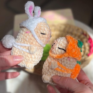 May include: Two crocheted bunny figures. One bunny is wearing a white hat with pink inner ears and a white tail. The other bunny is wearing an orange carrot hat. Both are beige with closed eyes.