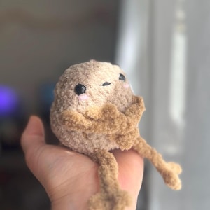 May include: A handmade plush toy shaped like a small, round creature. It has a light brown body and darker brown limbs. The toy features black eyes and pink cheeks, giving it a cute appearance. The plush is soft and fuzzy.