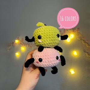 Puede incluir: Dos peluches de ganchillo apilados uno encima del otro. El juguete superior es de color verde lima y el inferior es rosa. Ambos tienen brazos y piernas negros, ojos negros y mejillas rosas. Un bocadillo rosa dice "16 COLORES".