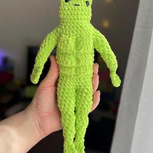 May include: A handmade, lime green crochet frog doll with black eyes. The frog has a textured, plush appearance, with arms and legs extended. The doll is held in a person's hand. The frog's body is segmented, and the head has a simple, rounded shape.