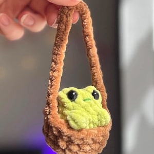 Könnte beinhalten: Handgehäkelter Frosch in einem braunen Korb. Der Frosch ist hellgrün mit schwarzen Augen und einem kleinen grünen Strich als Mund. Der Korb hat einen braunen Griff.