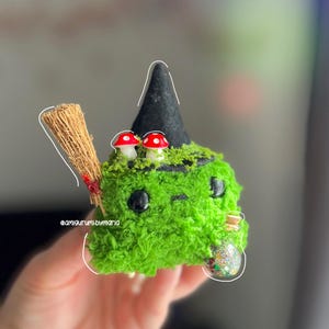 Puede incluir: Una bruja amigurumi verde caprichosa con un sombrero negro, acentos de hongos rojos y blancos y una pequeña escoba. La bruja tiene ojos negros y una pequeña botella de vidrio llena de brillantina. Hecho a mano.