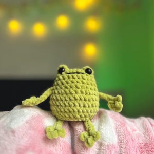 Op de afbeelding: Een handgemaakte, lichtgroene gehaakte kikker met zwarte ogen en uitgestrekte armen. De kikker zit op een roze en witte deken. De achtergrond is wazig met gele lichten en een groene muur.