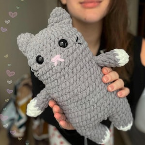 Könnte beinhalten: Ein handgefertigtes, graues Häkelkatzen-Plüschtier mit schwarzen Augen, einer rosa Nase und weißen Pfoten. Die Katze hat einen zwinkernden Gesichtsausdruck und wird in den Händen gehalten. Das Plüschtier ist ein weiches, kuscheliges Spielzeug.