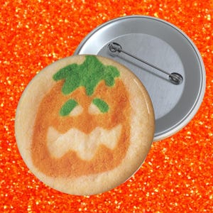 May include: A round pinback button featuring a printed image of a beige cookie shaped like a jack-o'-lantern with a green stem, green eyes, and a jagged white mouth. The back of the button is silver-colored metal with a horizontal pin closure.