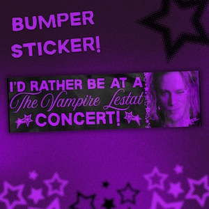 Könnte beinhalten: Ein rechteckiger Autoaufkleber mit schwarzem Hintergrund und violettem Text, der I'D RATHER BE AT A The Vampire Lestat CONCERT! lautet. Das Design enthält violette Sternmotive und ein Porträt einer Person mit langem Haar auf der rechten Seite.