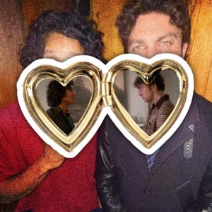 May include: A gold-toned heart-shaped locket featuring two open compartments. Each side of the locket displays a photograph of a person. The left side shows a person in a dark green shirt, and the right side shows a person in a patterned shirt.