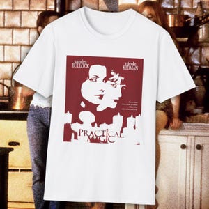 May include: A white short-sleeved t-shirt featuring a maroon graphic print of the movie poster for Practical Magic. The design includes the names Sandra Bullock and Nicole Kidman, the film title, and a silhouette of two faces above candles.