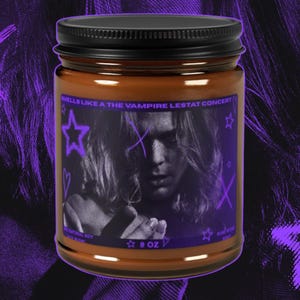 May include: A brown candle in a glass jar with a black lid. The label features a purple-toned portrait of a person with long hair, purple stars, and the text: SMELLS LIKE A THE VAMPIRE LESTAT CONCERT. The label also includes the text 9 OZ.