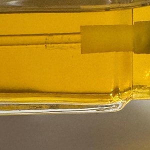 Puede incluir: Primer plano de una botella de vidrio llena de un l&iacute;quido amarillo dorado. La botella tiene forma rectangular con un borde ligeramente curvado. El l&iacute;quido es visible a trav&eacute;s del vidrio transparente y la botella parece estar sellada.