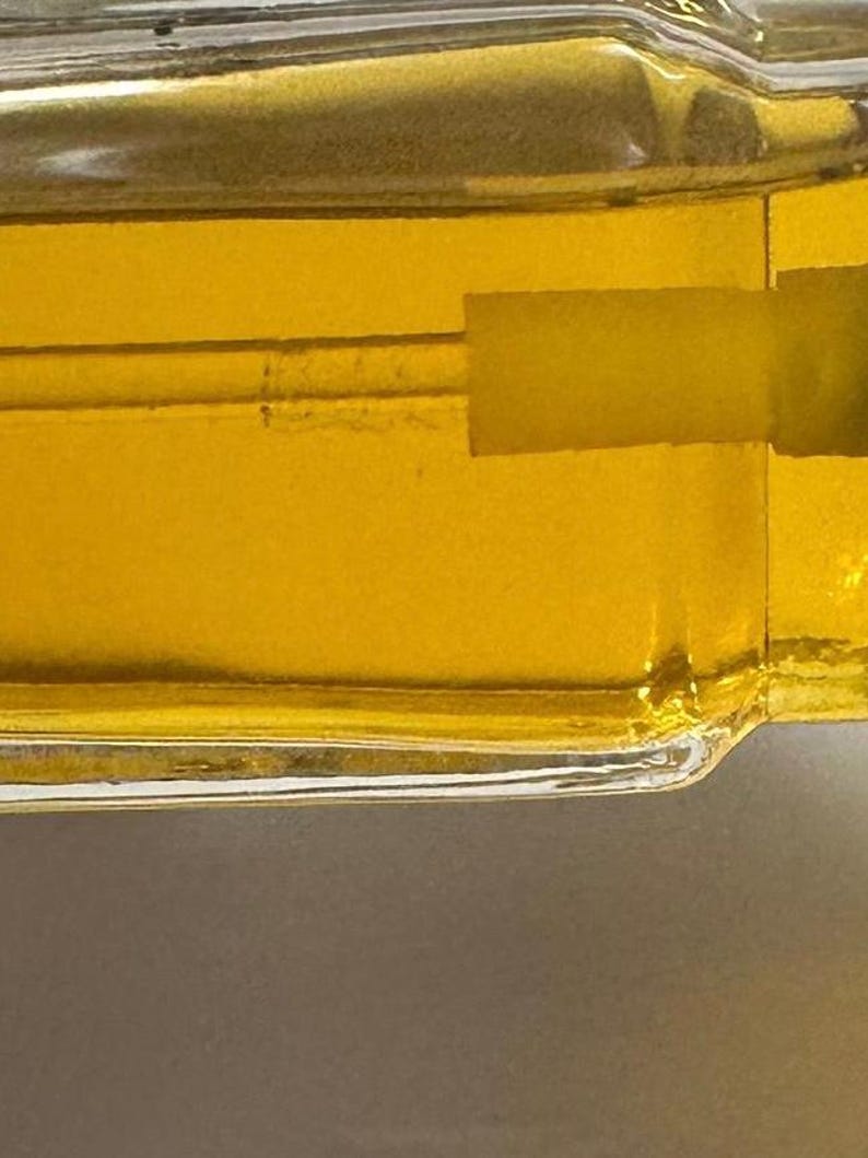 Puede incluir: Primer plano de una botella de vidrio llena de un l&iacute;quido amarillo dorado. La botella tiene forma rectangular con un borde ligeramente curvado. El l&iacute;quido es visible a trav&eacute;s del vidrio transparente y la botella parece estar sellada.