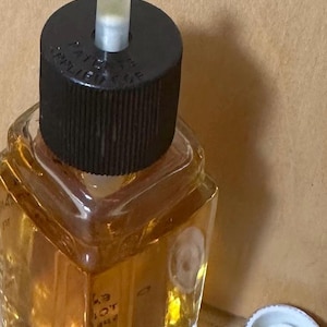 Puede incluir: Un frasco de perfume vintage con una tapa negra y un aplicador de vidrio transparente. El frasco est&aacute; lleno de un l&iacute;quido de color dorado. La tapa tiene texto. Una tapa blanca est&aacute; al lado del frasco.