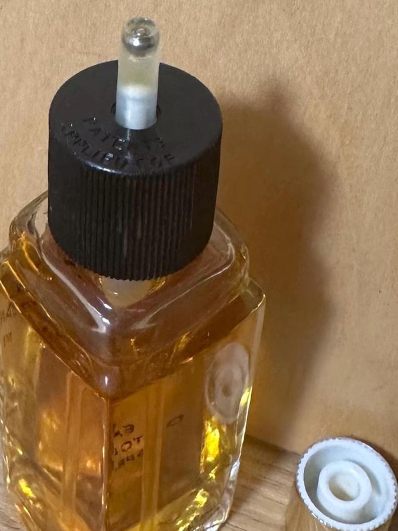 Puede incluir: Un frasco de perfume vintage con una tapa negra y un aplicador de vidrio transparente. El frasco est&aacute; lleno de un l&iacute;quido de color dorado. La tapa tiene texto. Una tapa blanca est&aacute; al lado del frasco.