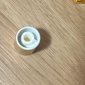 Puede incluir: Una tapa de botella plateada con interior blanco y una abertura central. La tapa tiene un borde estriado y un dise&ntilde;o circular. La tapa est&aacute; sobre una superficie de madera.