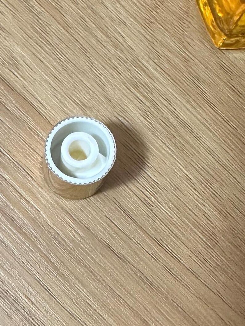 Puede incluir: Una tapa de botella plateada con interior blanco y una abertura central. La tapa tiene un borde estriado y un dise&ntilde;o circular. La tapa est&aacute; sobre una superficie de madera.