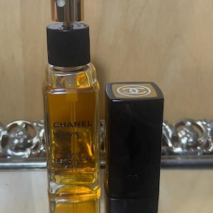 Puede incluir: Un frasco de Chanel No. 5 Eau de Toilette Spray con tap&oacute;n dorado y base negra. El frasco es rectangular y est&aacute; lleno de l&iacute;quido dorado. Junto a &eacute;l, un estuche de l&aacute;piz labial rectangular negro con un logotipo dorado.