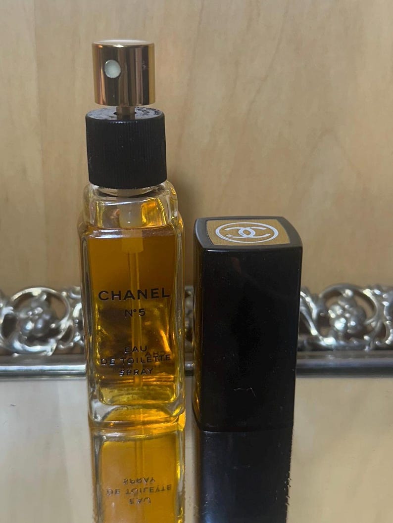 Puede incluir: Un frasco de Chanel No. 5 Eau de Toilette Spray con tap&oacute;n dorado y base negra. El frasco es rectangular y est&aacute; lleno de l&iacute;quido dorado. Junto a &eacute;l, un estuche de l&aacute;piz labial rectangular negro con un logotipo dorado.