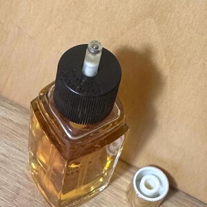 Puede incluir: Un peque&ntilde;o frasco de perfume de vidrio transparente con una tapa negra y una tapa dorada separada. La botella est&aacute; llena de un l&iacute;quido dorado. La botella tiene una forma geom&eacute;trica y est&aacute; sobre una superficie de madera.