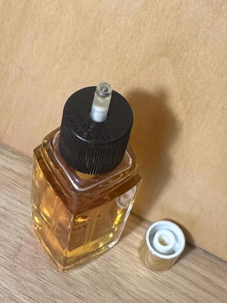 Puede incluir: Un peque&ntilde;o frasco de perfume de vidrio transparente con una tapa negra y una tapa dorada separada. La botella est&aacute; llena de un l&iacute;quido dorado. La botella tiene una forma geom&eacute;trica y est&aacute; sobre una superficie de madera.