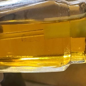 Puede incluir: Una botella de vidrio transparente llena de l&iacute;quido dorado. La botella tiene forma rectangular con bordes redondeados. El l&iacute;quido en el interior es de color &aacute;mbar claro. La botella tiene una etiqueta con texto.