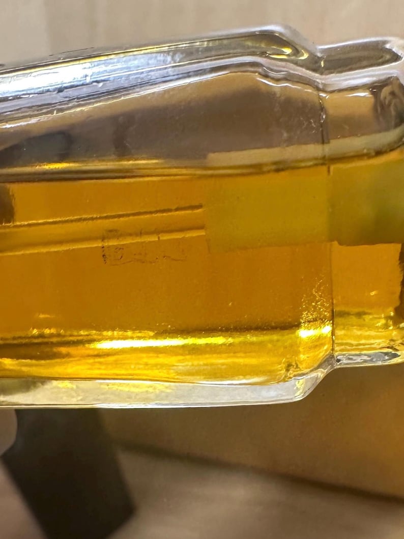 Puede incluir: Una botella de vidrio transparente llena de l&iacute;quido dorado. La botella tiene forma rectangular con bordes redondeados. El l&iacute;quido en el interior es de color &aacute;mbar claro. La botella tiene una etiqueta con texto.