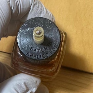 Puede incluir: Una peque&ntilde;a botella cuadrada de vidrio con una tapa oscura. La tapa tiene las palabras "PATENTS APPLIED FOR" alrededor del borde. La botella contiene un l&iacute;quido marr&oacute;n claro. La botella es sostenida por una mano enguantada.