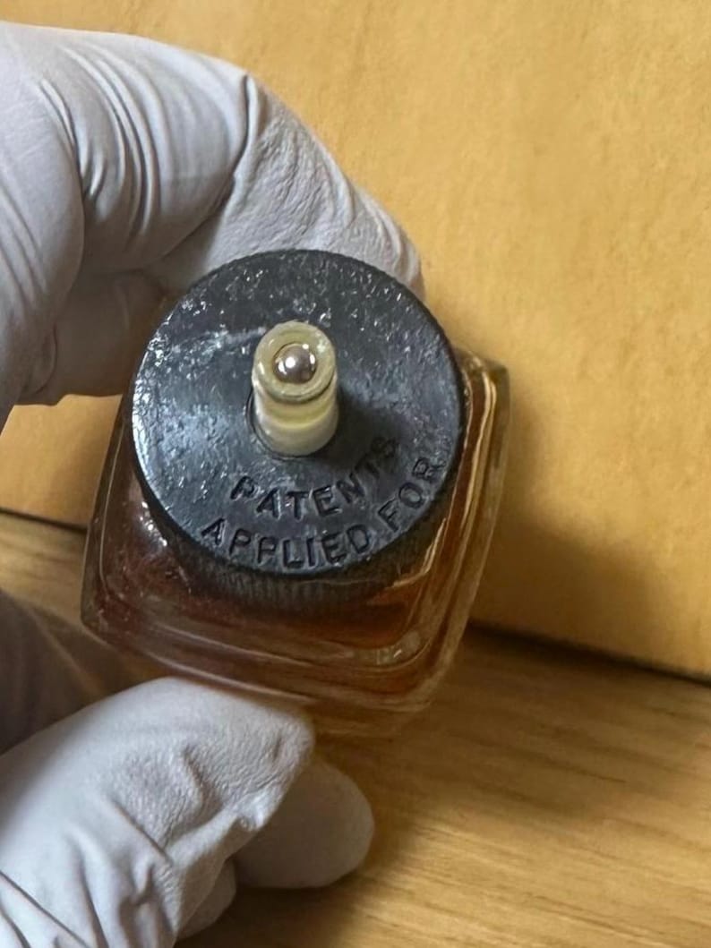 Puede incluir: Una peque&ntilde;a botella cuadrada de vidrio con una tapa oscura. La tapa tiene las palabras "PATENTS APPLIED FOR" alrededor del borde. La botella contiene un l&iacute;quido marr&oacute;n claro. La botella es sostenida por una mano enguantada.