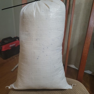 Könnte beinhalten: Ein großer weißer Jutesack mit einem schwarzen Kordelverschluss. Der Sack ist mit einer weißen Substanz gefüllt und steht auf einem braunen Stuhl.