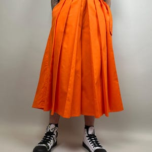 Può includere: Una gonna plissettata di colore arancione brillante con una cintura larga. La gonna è lunga fino al ginocchio e ha una silhouette fluida.
