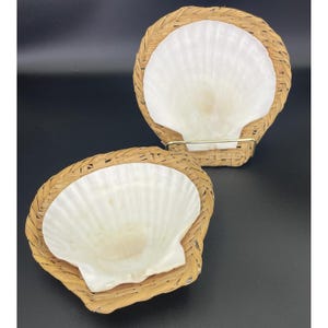 May include: Two white scallop shells are set in woven wicker baskets. The baskets have a circular shape with a woven border. The shells are a creamy white color and have a textured surface. The background is black.