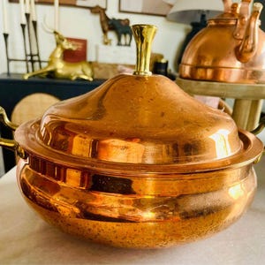 May include: A copper pot with a lid and two handles. The pot has a shiny, polished finish and a brass knob on the lid.