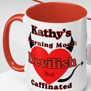 May include: White ceramic coffee mug with a red rim. The mug has a black and red graphic with the text "Kathy's Morning Mood: Devilish But Caffinated".