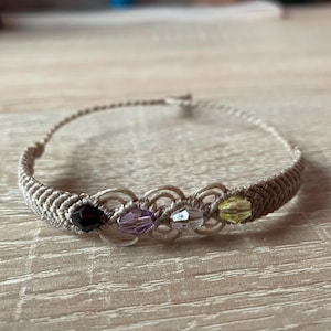 Könnte beinhalten: Ein beigefarbenes Makramee-Armband mit vier kleinen Glasperlen in den Farben Lila, Klar und Gelb.